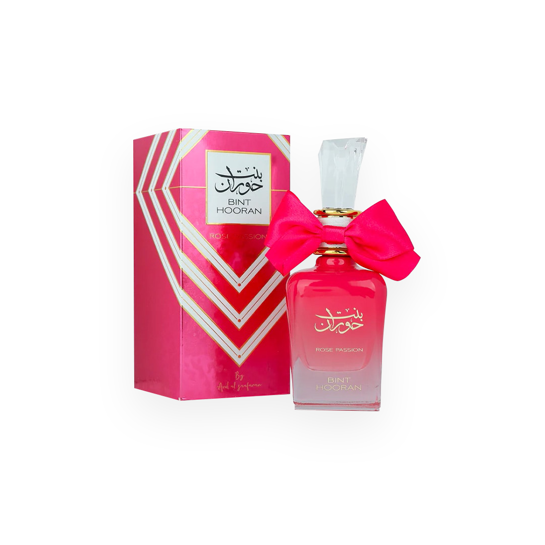 Une bouteille de parfum rose vif appelée "Bint Hooran Rose Passion" de la marque Ard Al Zaafaran, présentée dans un flacon élégant avec un grand nœud rose attaché autour du col et un bouchon transparent. La boîte d'emballage, assortie à la couleur du flacon, affiche des motifs géométriques avec des accents dorés et le nom du produit en arabe et en anglais.