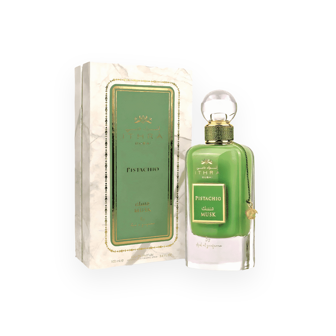 Un flacon de parfum vert intense appelé "Pistachio Musk" de la marque Ithra Dubai par Ard Al Zaafaran. Le flacon raffiné est surmonté d’un bouchon sphérique transparent et décoré d’un cordon doré avec un petit pendentif assorti. L’emballage présente un design chic avec un fond marbré blanc, des accents dorés et une étiquette verte élégante au centre, mettant en avant le nom du parfum en arabe et en anglais.