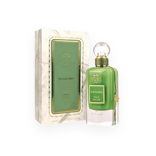 Un flacon de parfum vert intense appelé "Pistachio Musk" de la marque Ithra Dubai par Ard Al Zaafaran. Le flacon raffiné est surmonté d’un bouchon sphérique transparent et décoré d’un cordon doré avec un petit pendentif assorti. L’emballage présente un design chic avec un fond marbré blanc, des accents dorés et une étiquette verte élégante au centre, mettant en avant le nom du parfum en arabe et en anglais.